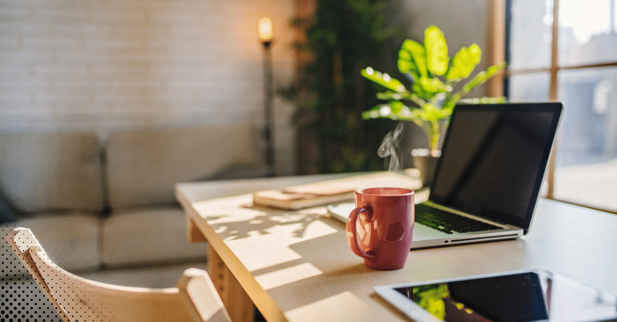 Cozy home workspace with a laptop, tablet, and steaming mug on a desk.